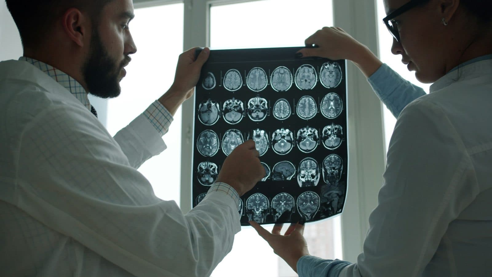 Two doctors hold up a sheet of brain MRI scans against a bright window to review them.