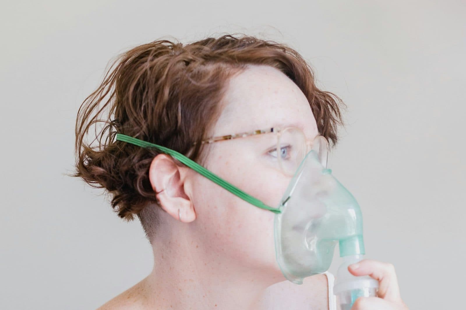 A woman with short curly hair holds a clear nebulizer mask over her nose and mouth.
