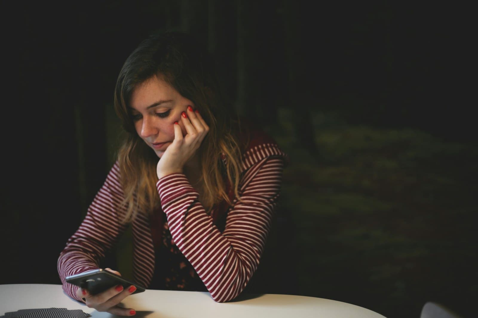 A woman rests her chin in her hand as she looks at her phone at a table in a dark room.