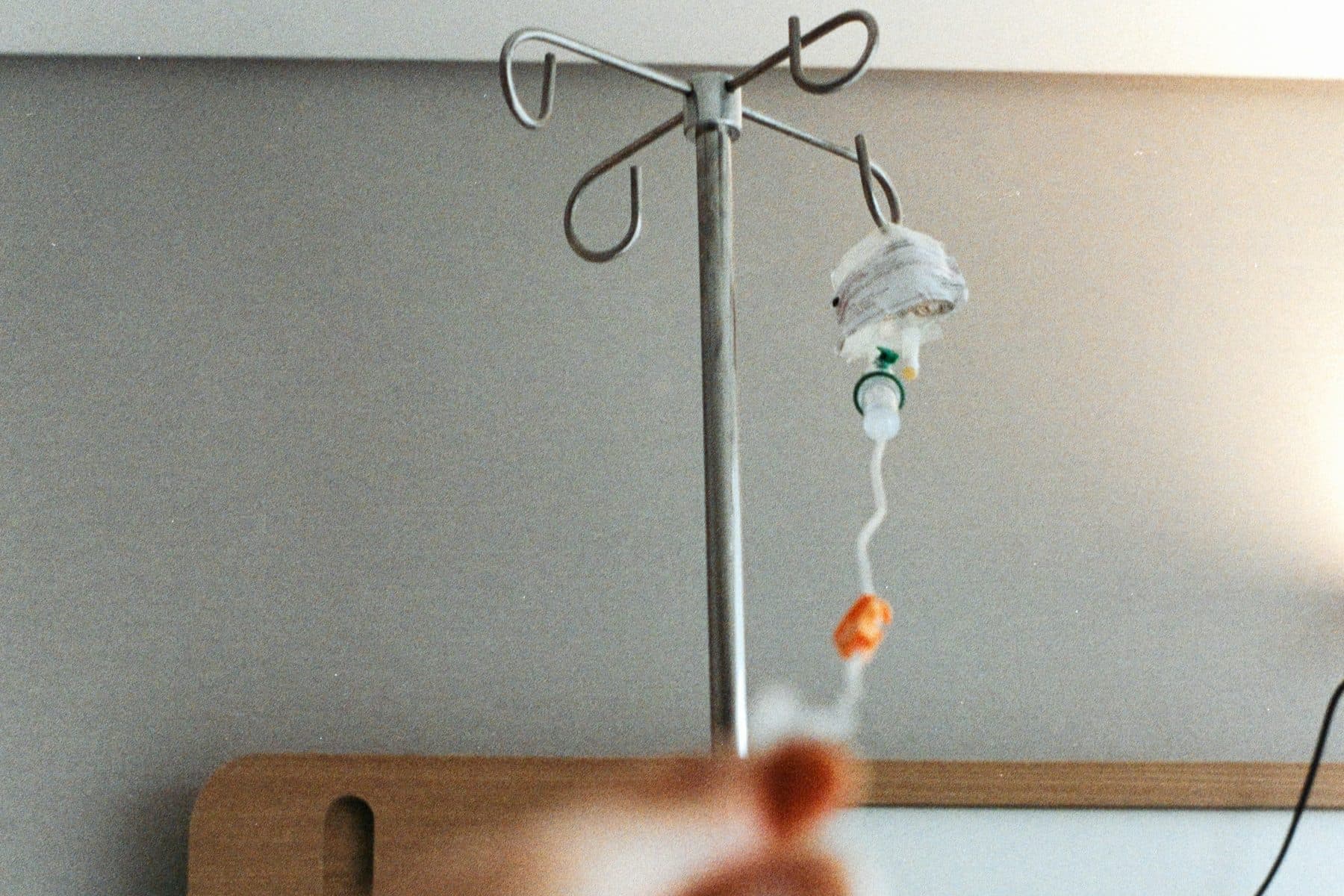 A hand reaches toward an IV pole with an infusion line hanging from it in a hospital room.