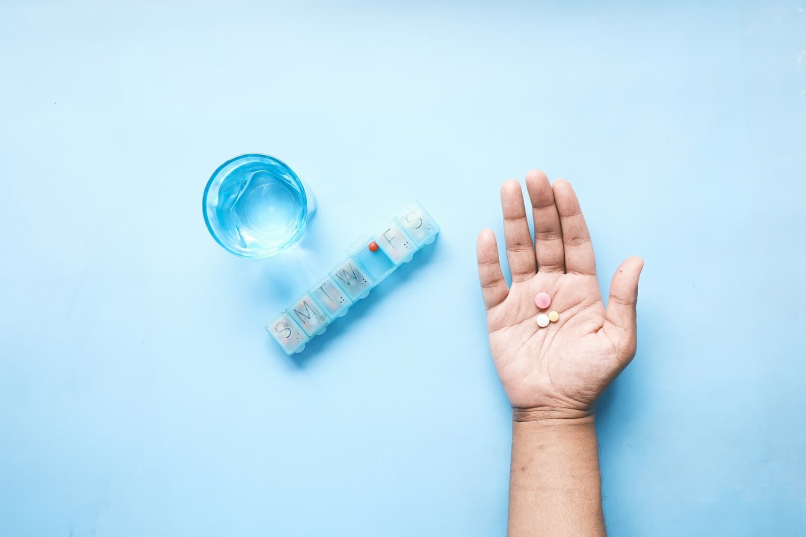 An open hand holding pills next to a weekly pill organizer and a glass of water on a light blue surface.