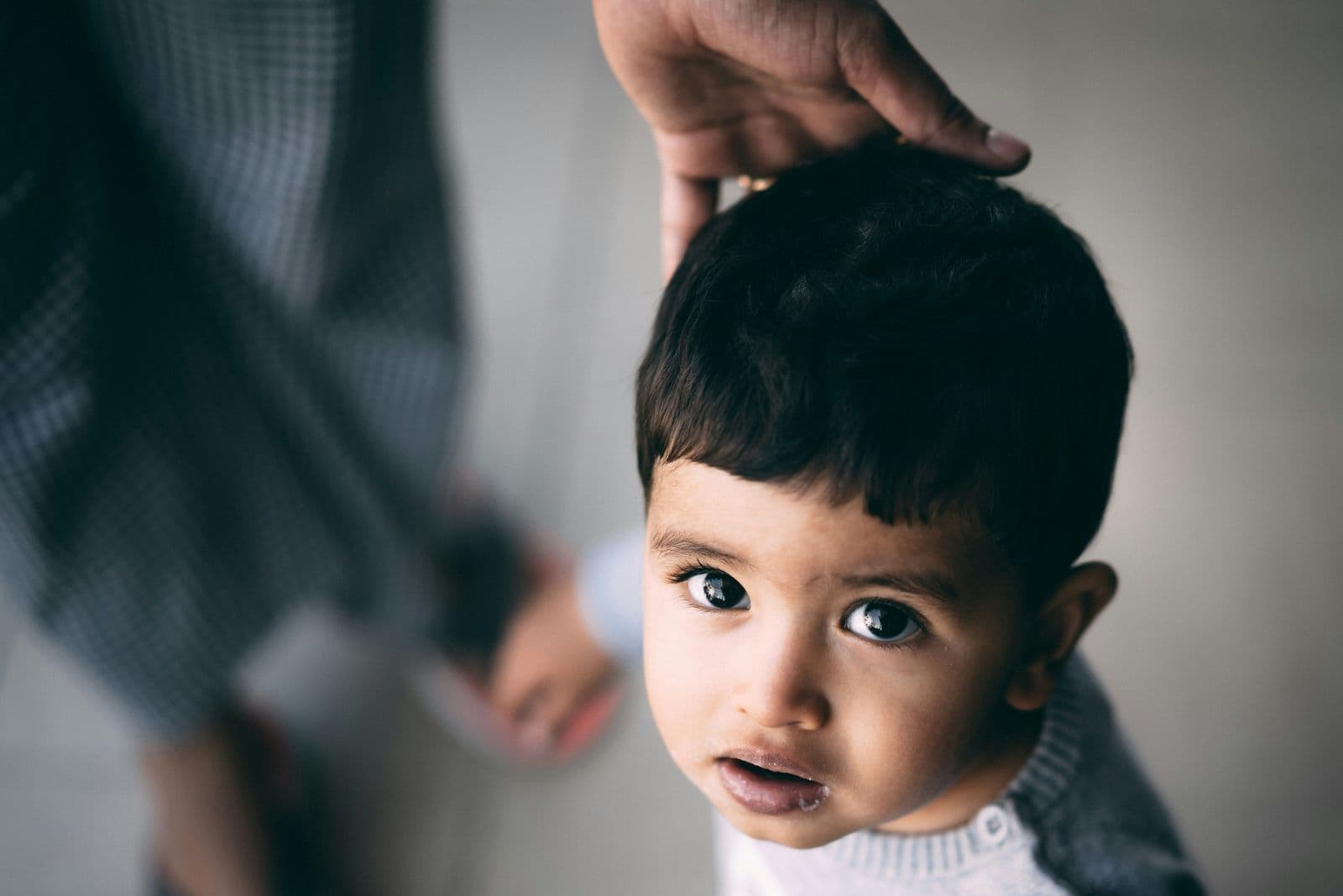 A parent rests a hand on a toddler's head as the little boy looks up at the camera.