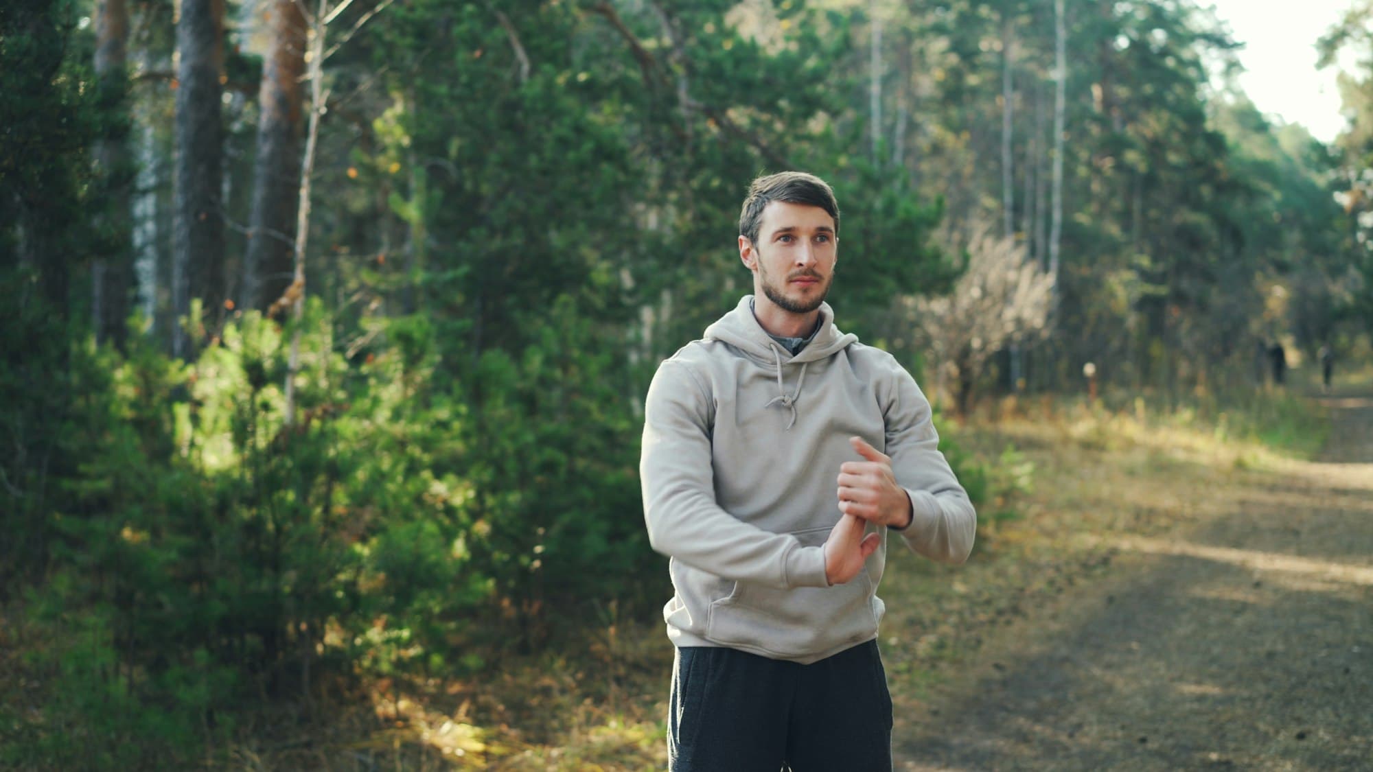 A young man in a grey hoodie stretches his arms on a sunlit forest path before a run.