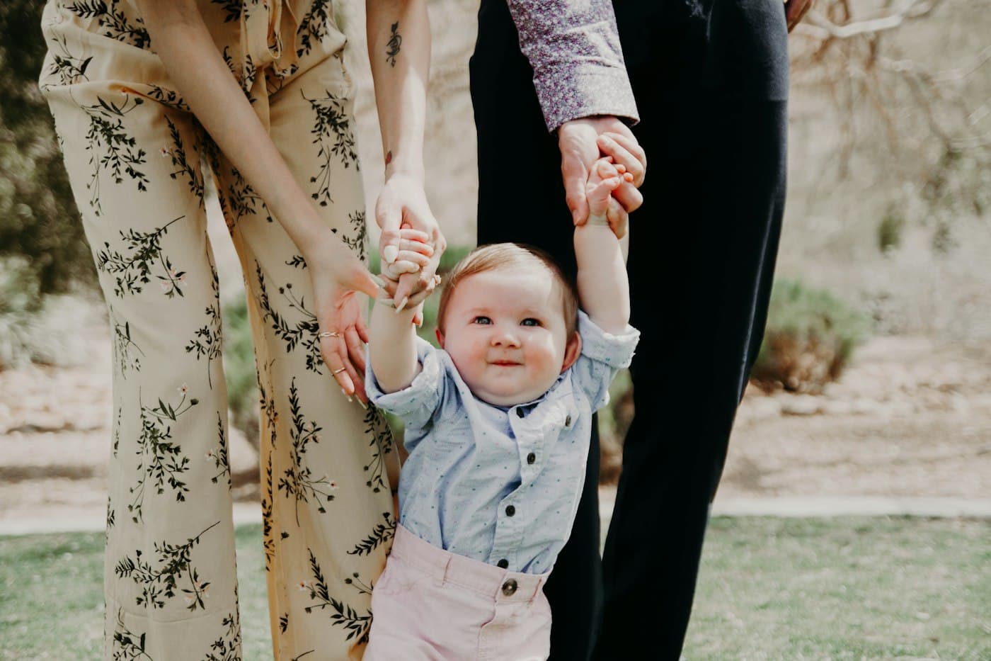 A toddler walking across grass while holding a parent's hand on each side.