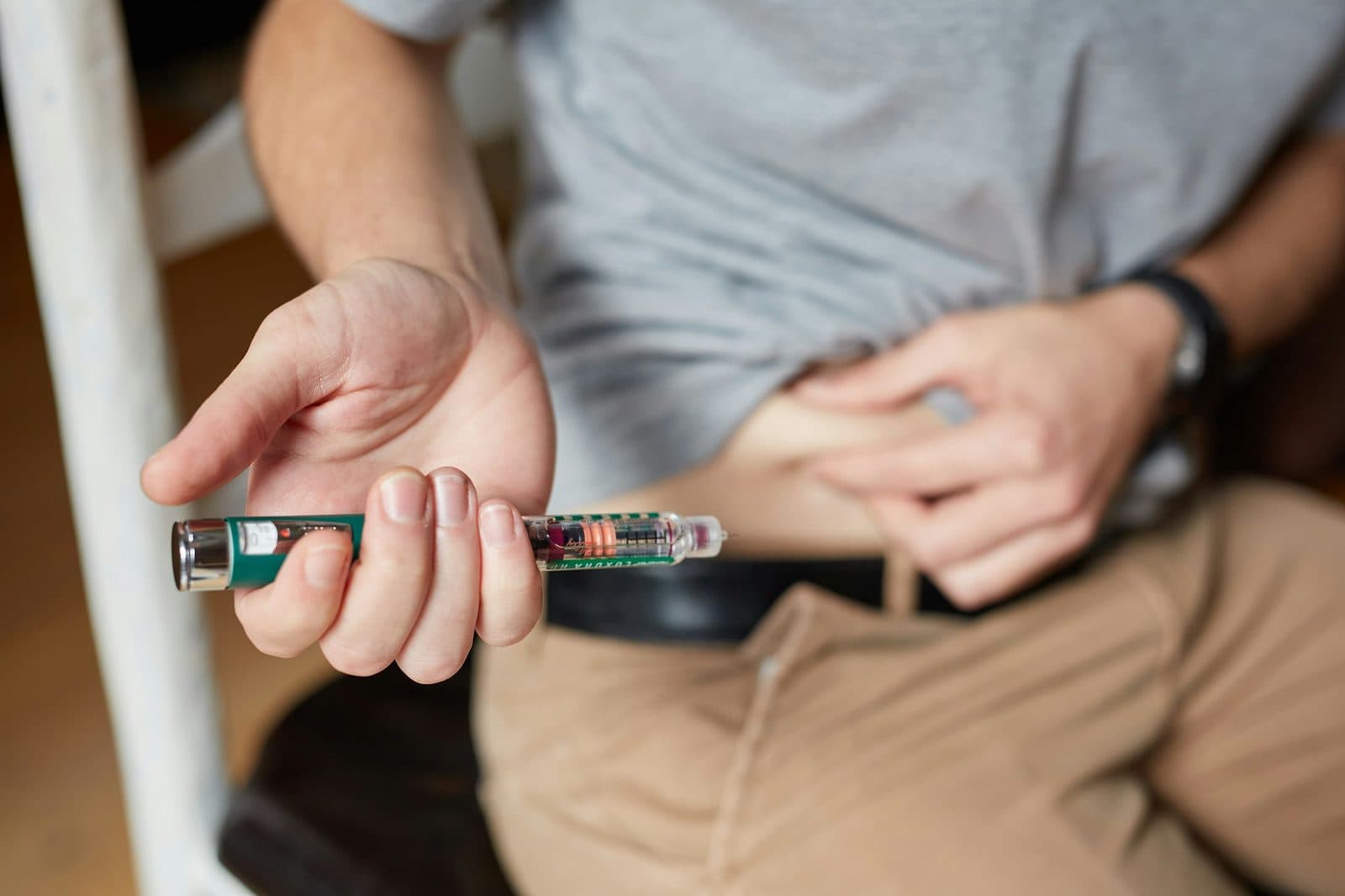 A man in a grey T-shirt holds an injection pen near his stomach, about to give himself a dose at home.