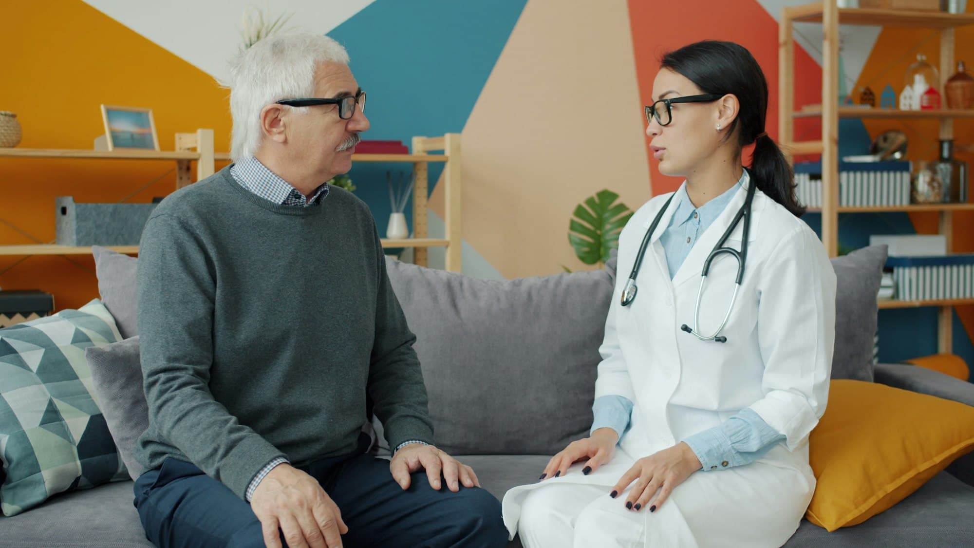 A doctor in a white coat talks with an older man with grey hair as they sit together on a couch.
