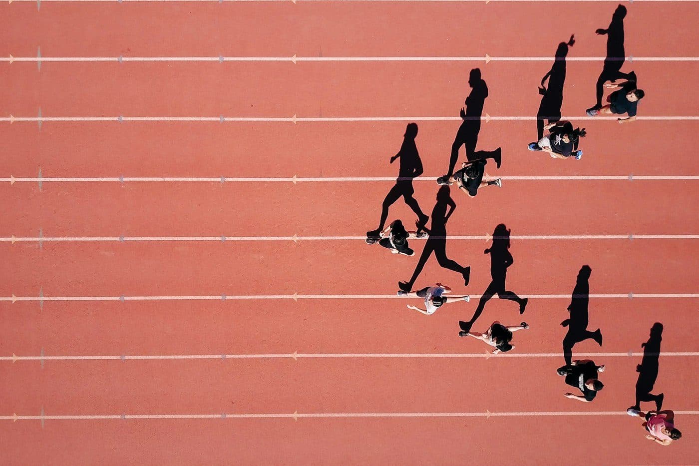 Overhead view of a group of runners on a red track, with one runner falling behind the pack.