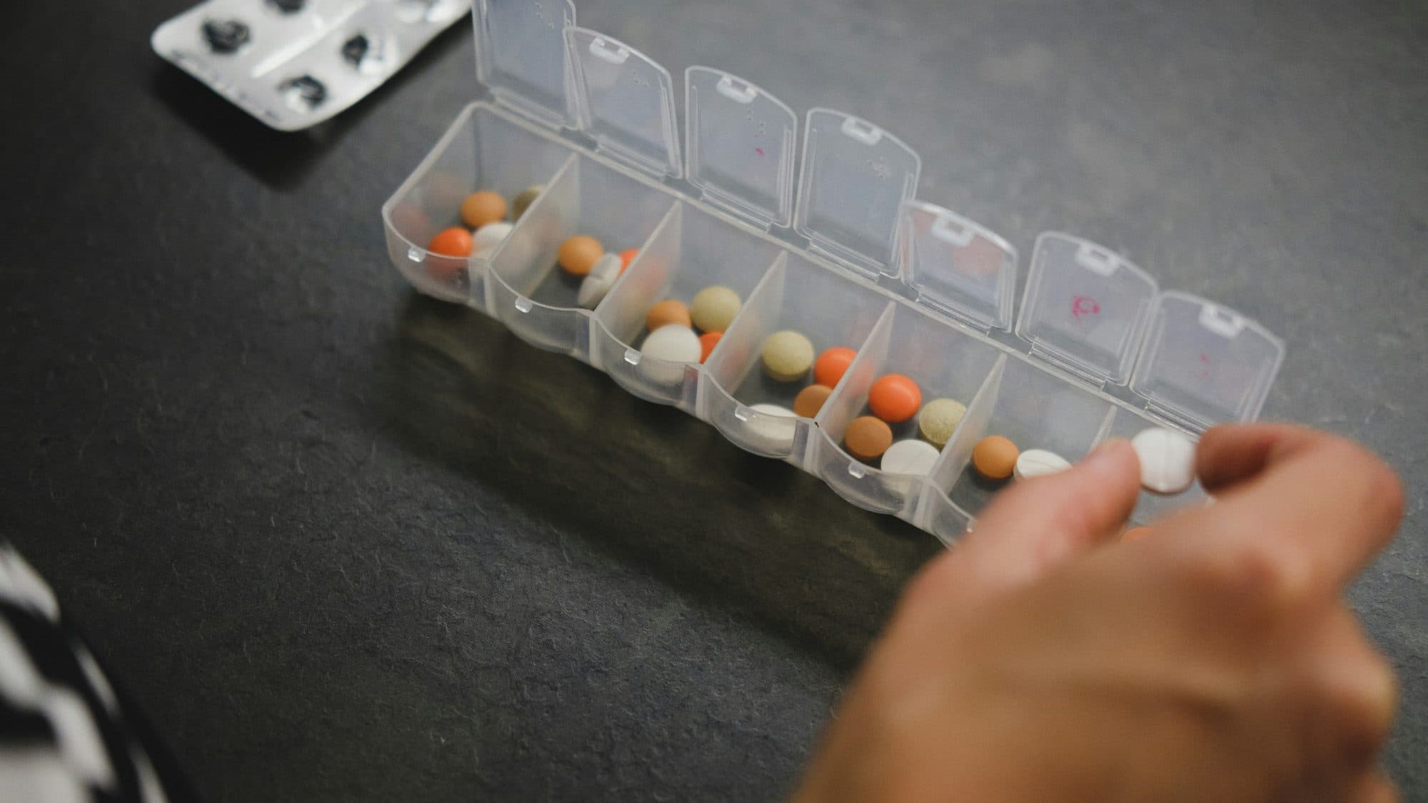 A hand takes a pill from a weekly pill organizer on a dark table, next to a blister pack.