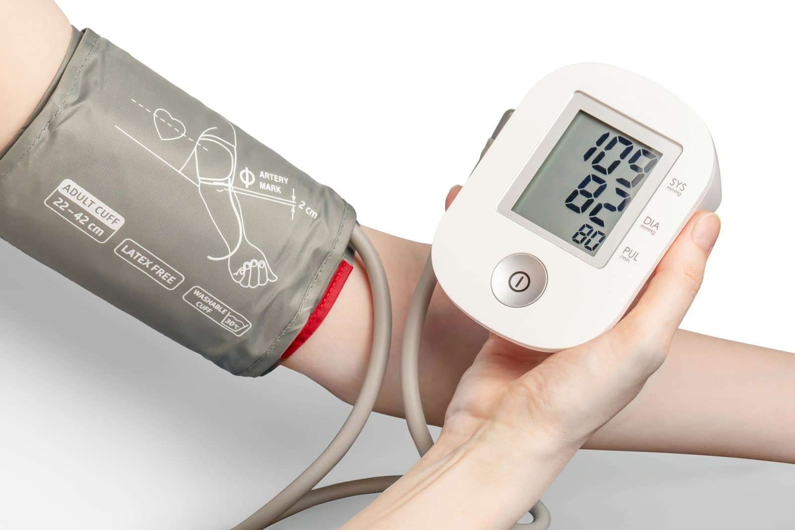 A hand holds a digital blood pressure monitor while a grey cuff is wrapped around the upper arm.