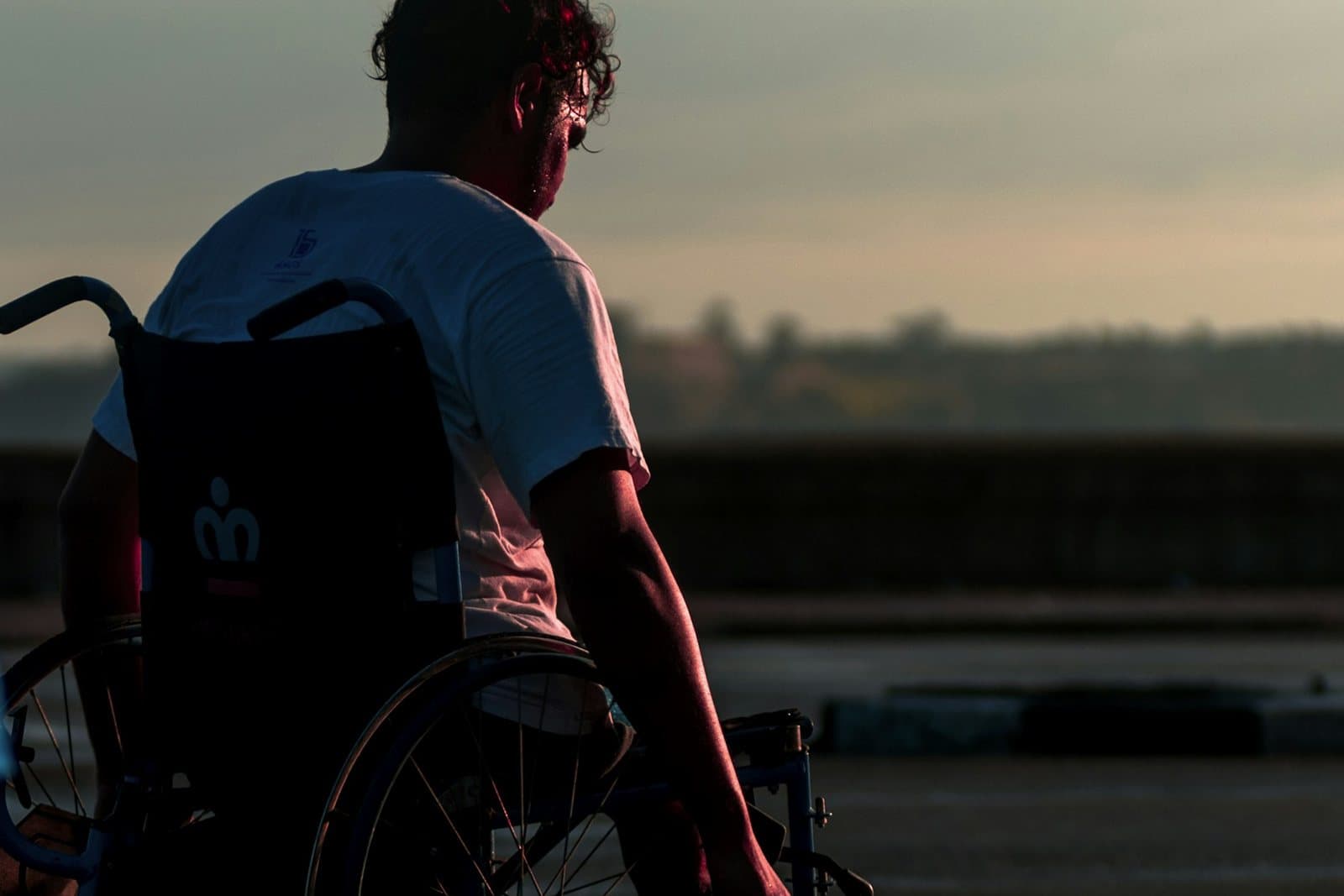 A young man in a wheelchair at sunset, seen from behind with one hand on the wheel rim.
