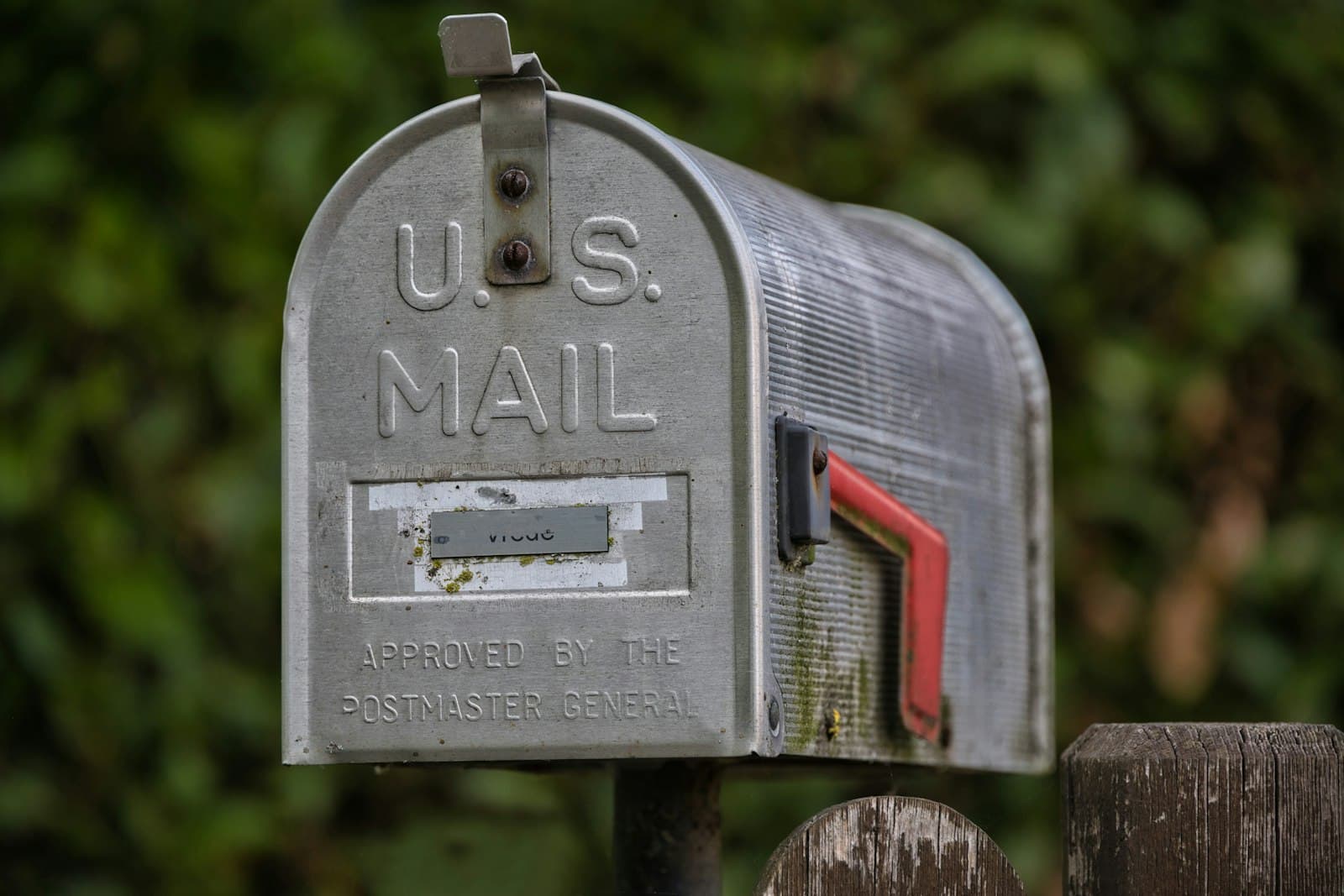 A weathered U.S. Mail mailbox with its red flag down, mounted beside a wooden fence post.
