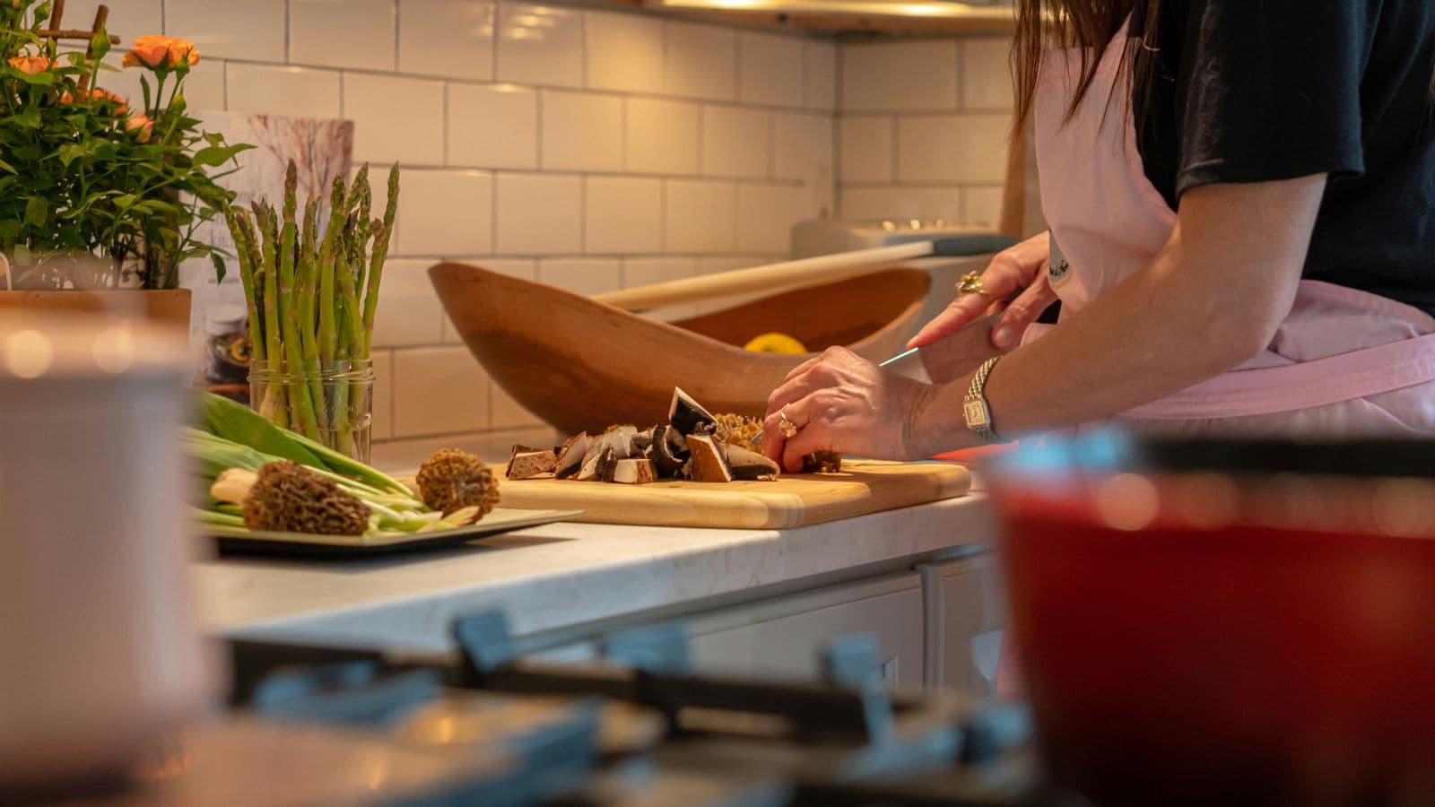 A woman in an apron chops mushrooms on a wooden board in a home kitchen, with asparagus and fresh vegetables on the counter.