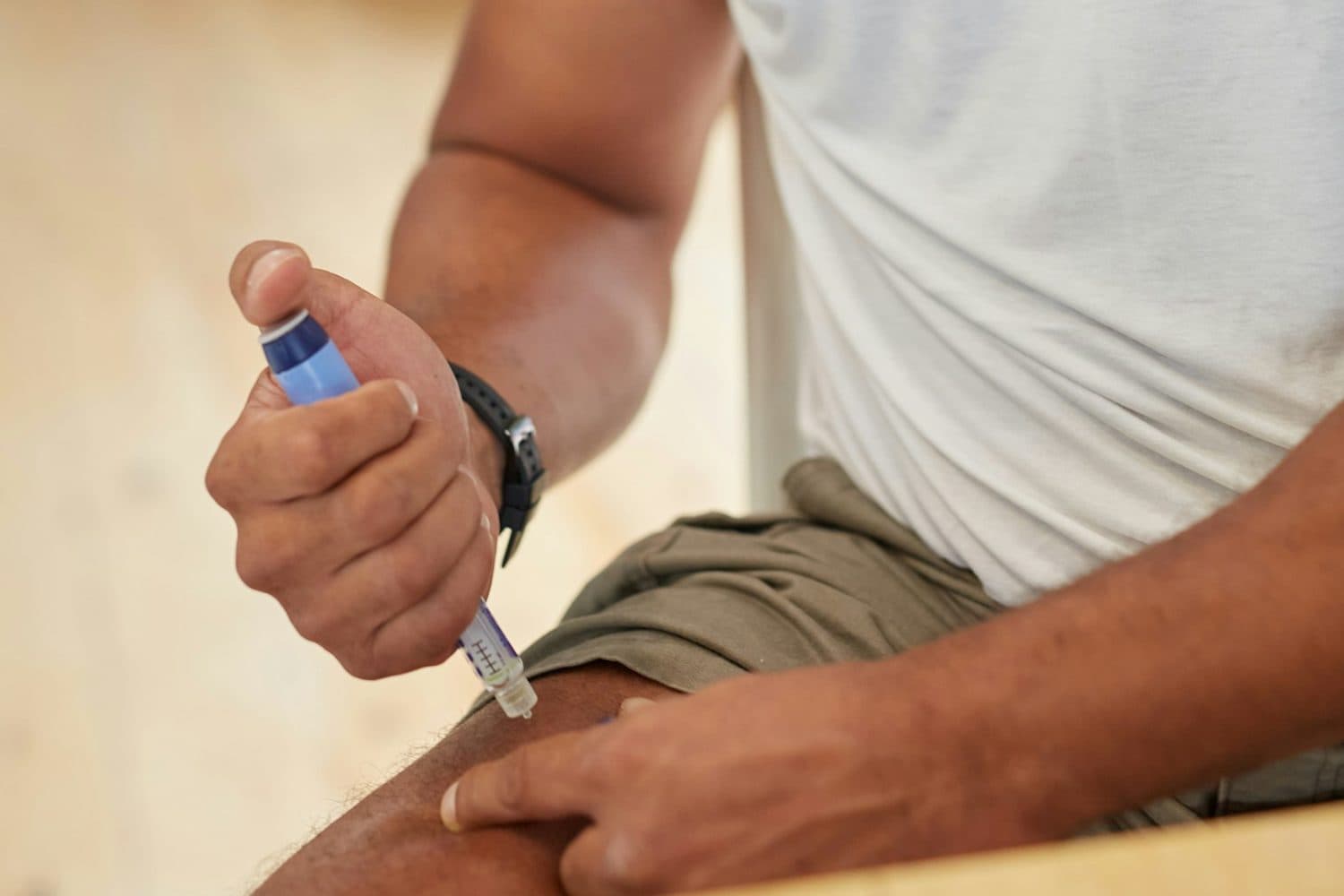 A man presses a pen injector into the front of his thigh while seated at home in a white T-shirt.