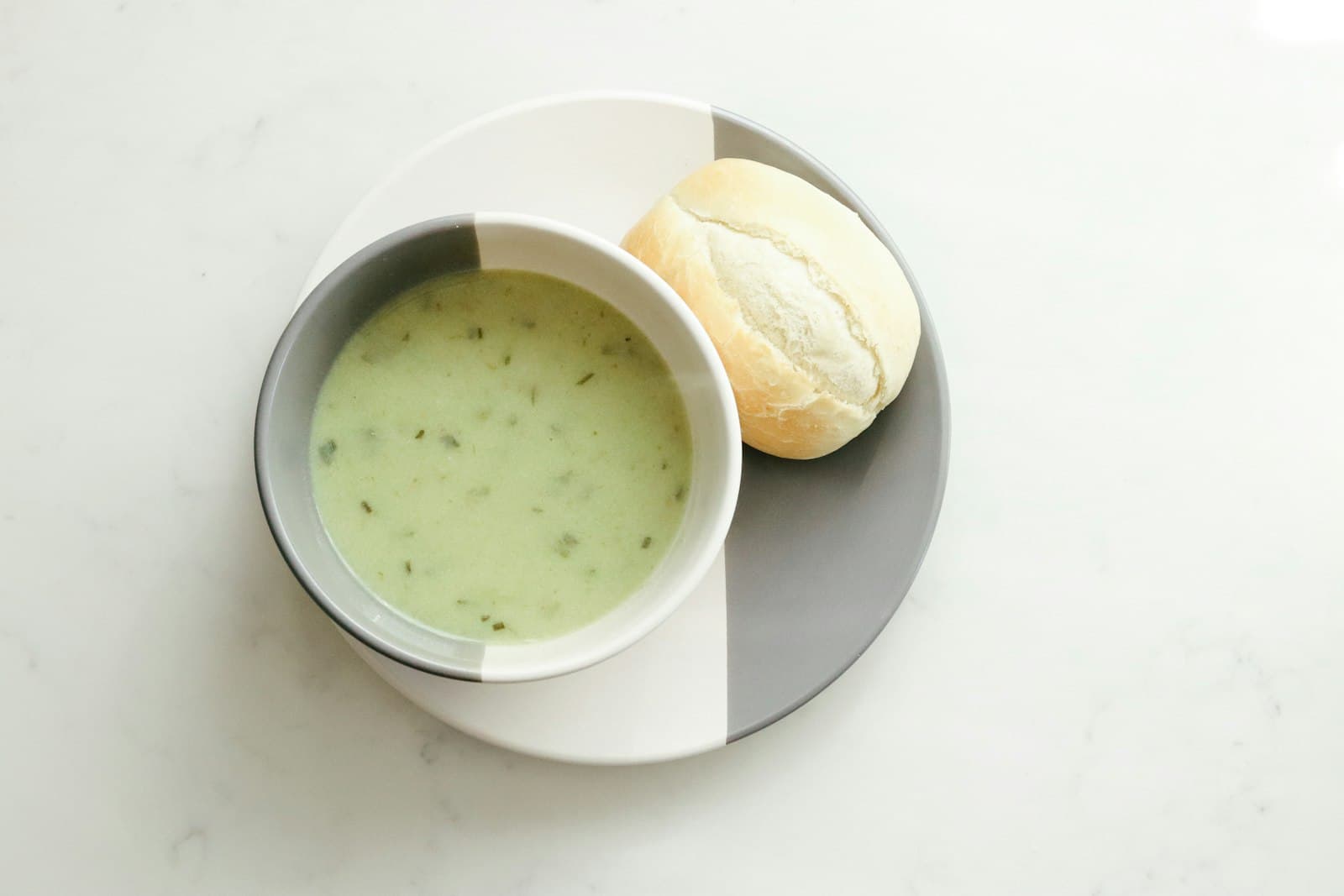 A bowl of creamy soup next to a small bread roll on a white table, seen from above.