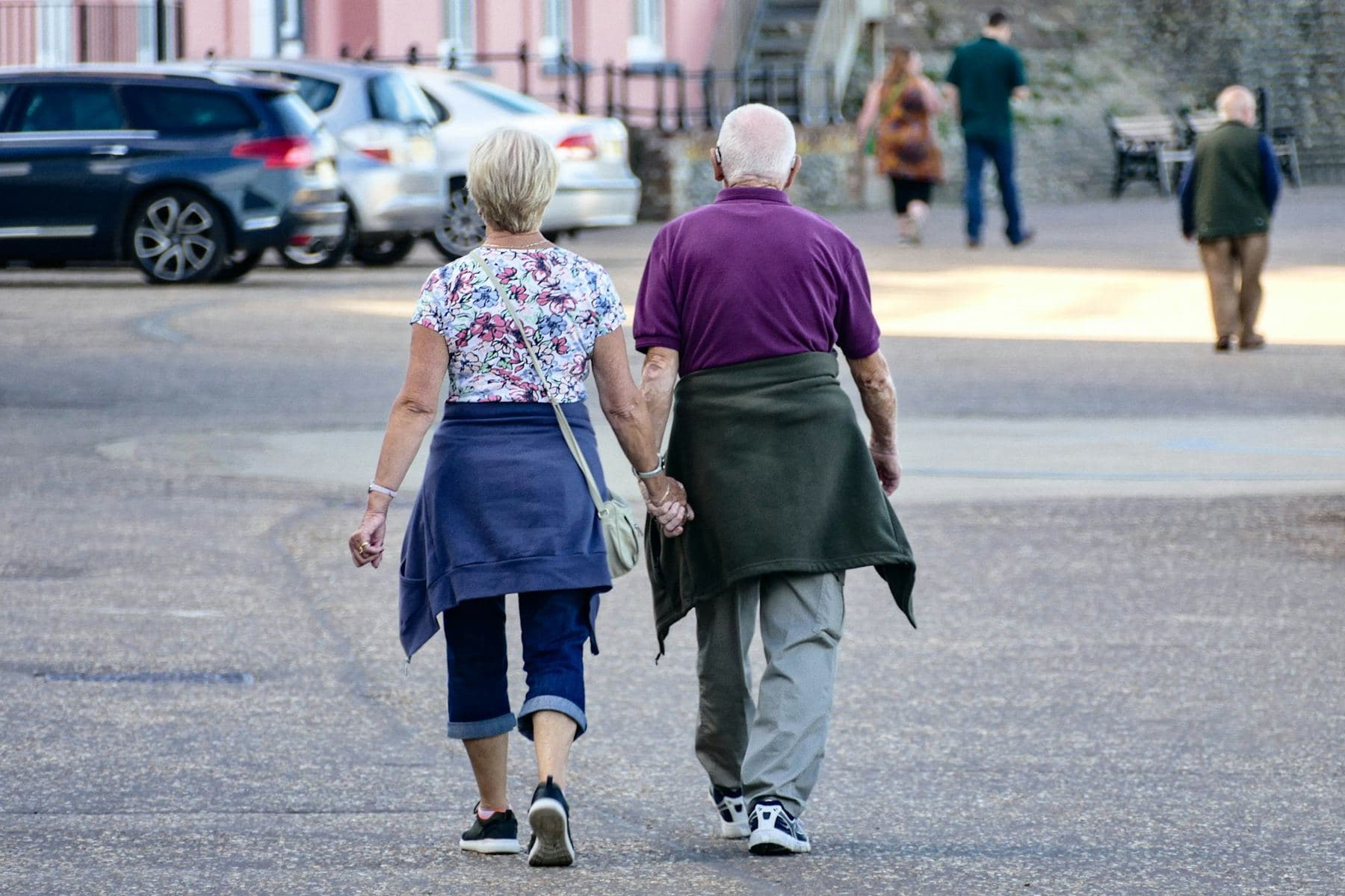 An older couple holds hands as they walk across a town square, seen from behind.
