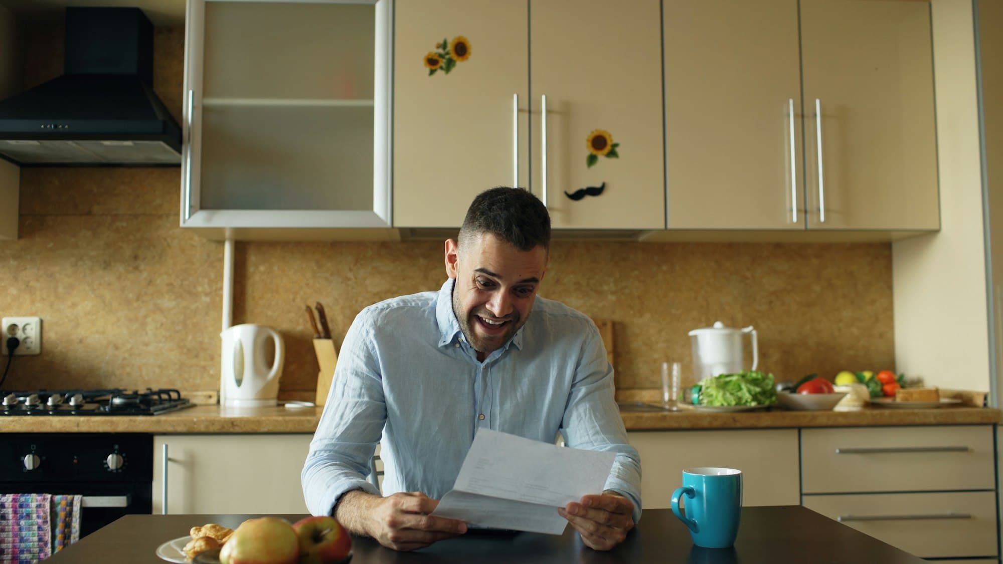 A man in a blue shirt reads a letter at his kitchen table, mouth open in surprise, next to a blue mug and a plate of fruit.