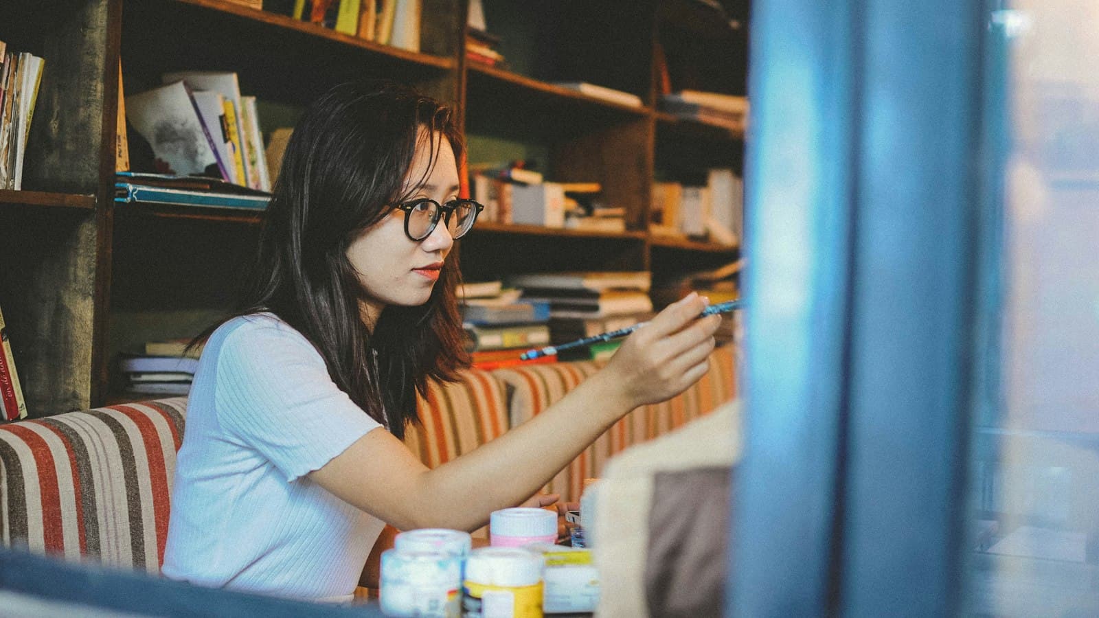 A woman in glasses paints with a brush at a table in front of tall bookshelves, with small jars of paint in the foreground.