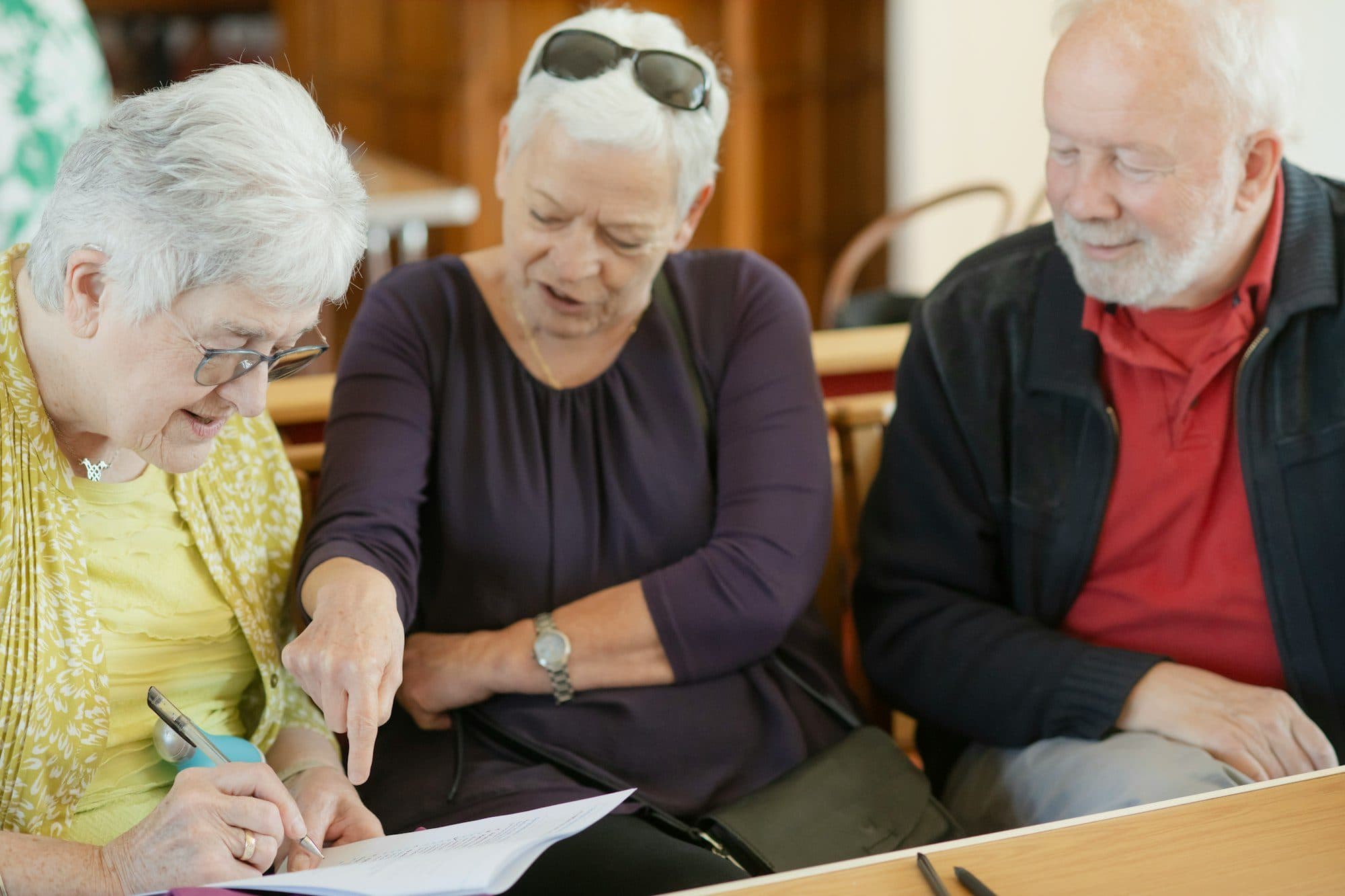 Three older adults at a table, one pointing at a form while another fills it in.