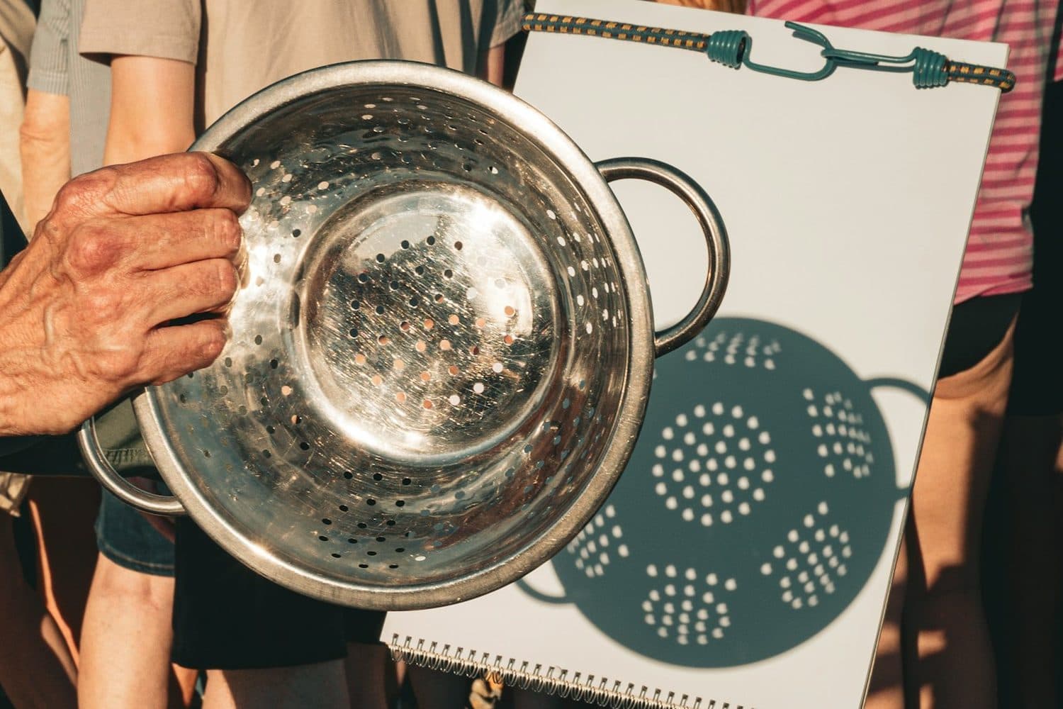 An older hand holds a metal colander up to the sun, casting a shadow of tiny holes onto a white board.