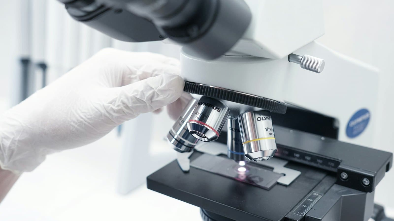 A gloved hand turns the lenses of a microscope over a glass tissue slide in a lab.