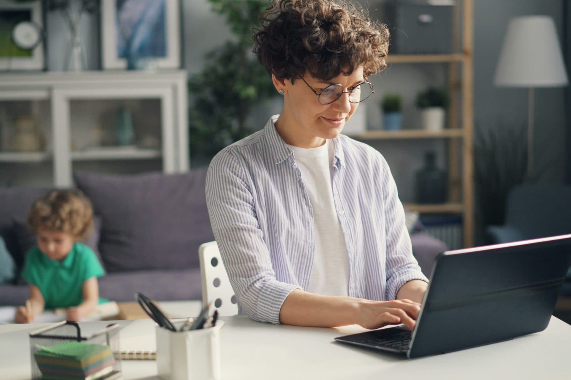 A mother types at a laptop on her dining table while her young son draws on the couch behind her.