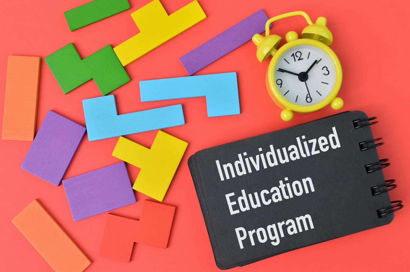 A black spiral notebook with the words Individualized Education Program in white lettering, surrounded by brightly colored wooden block pieces and a yellow alarm clock on a coral background, illustrating the planning, advocacy, and timing involved in setting up an IEP or 504 plan