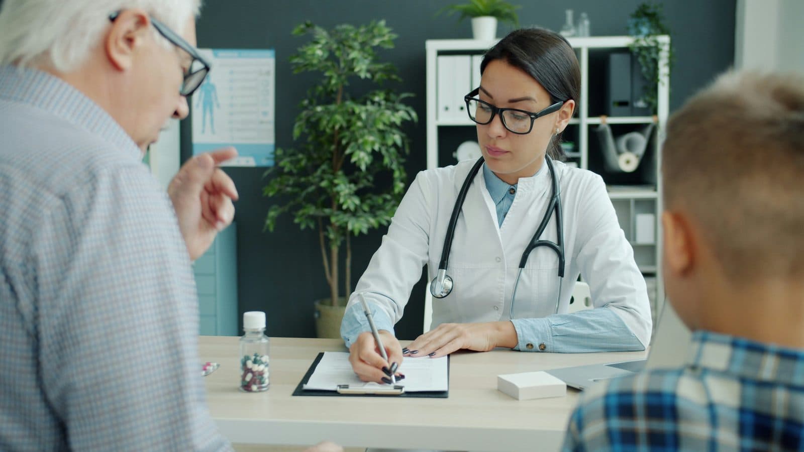 A doctor takes notes at her desk while an older man and a boy talk with her during a clinic visit.