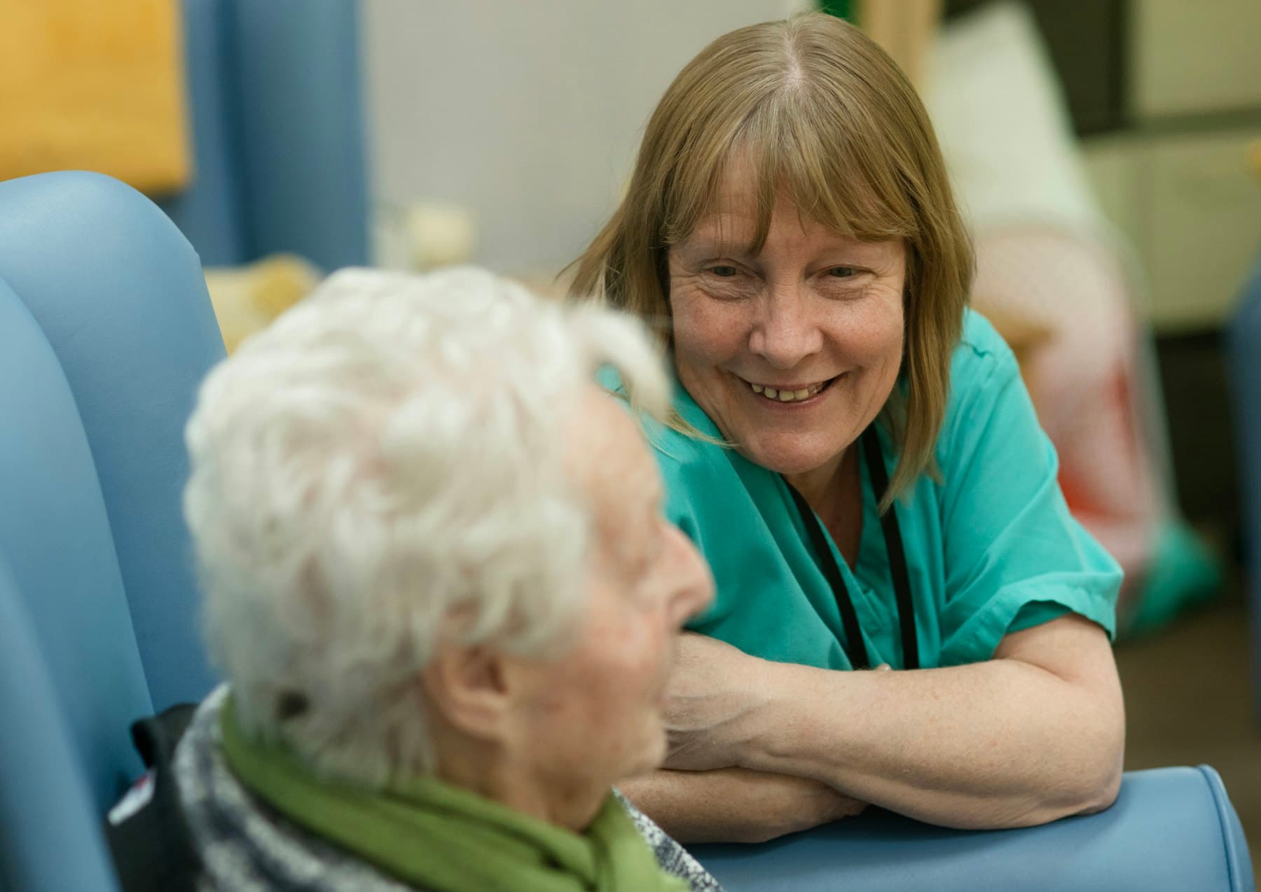 A caregiver in green scrubs leaning toward an older woman