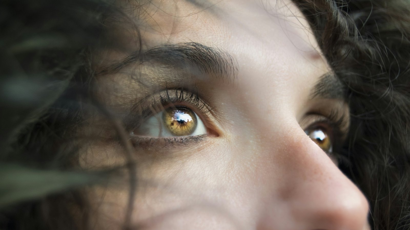 Close-up of a woman's hazel eyes looking upward, dark curls blurred around her face.