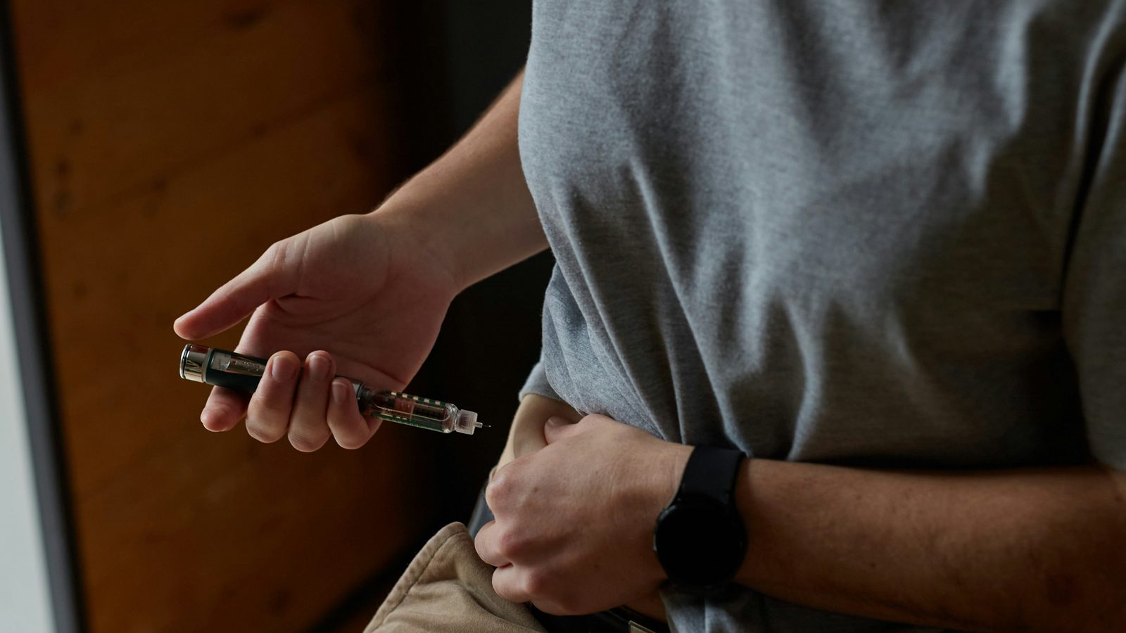 A person in a gray T-shirt pinches skin at the waist and holds a prefilled injection pen ready to inject at home.