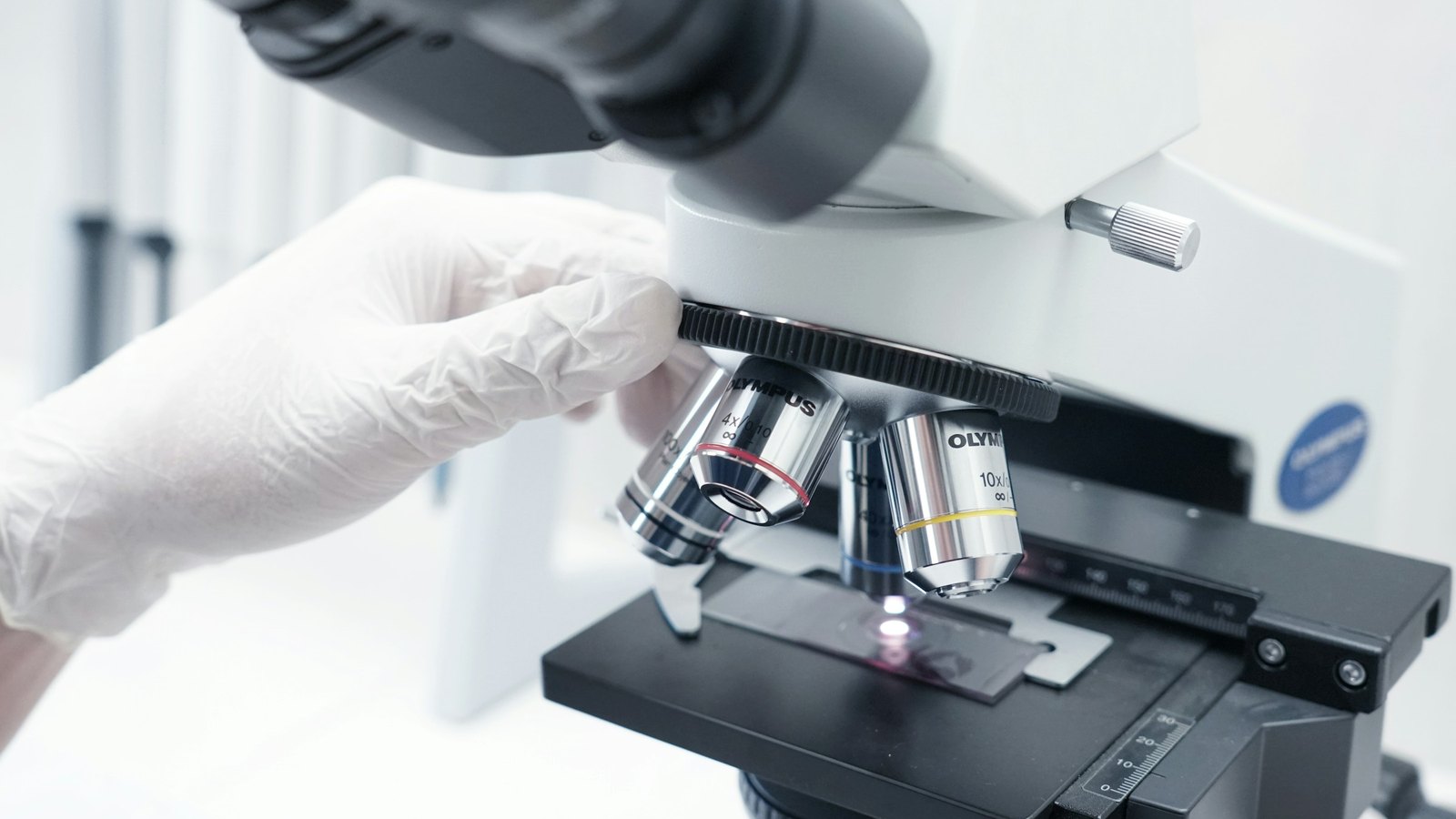 A gloved hand turns the lenses of a microscope over a glass tissue slide in a lab.